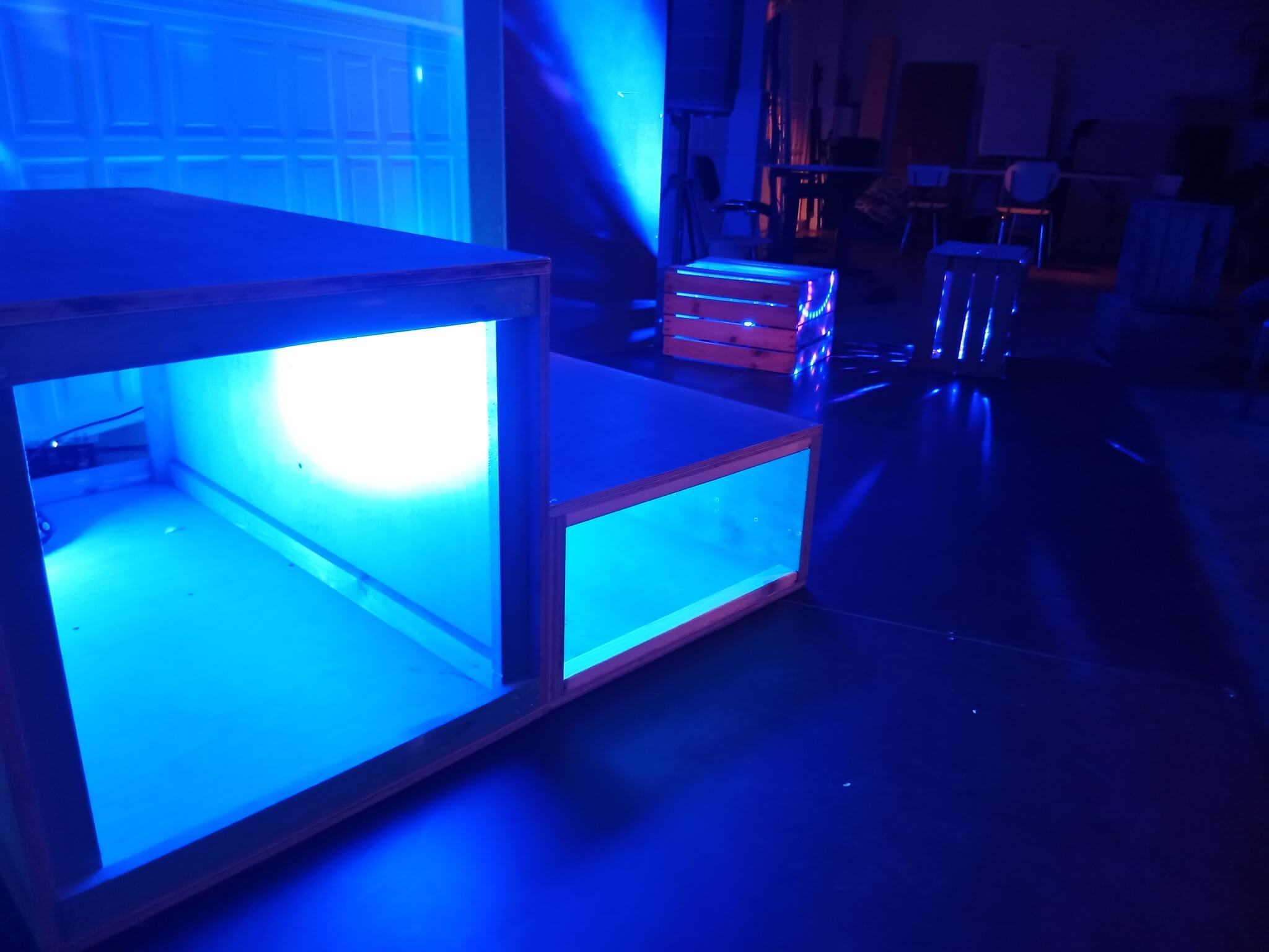 Photographie de la SALLE lors d'une installation lumineuse. Des blocs creus et des palettes de bois sont dans l'ombre, éclairées par de fortes lumières bleues.
